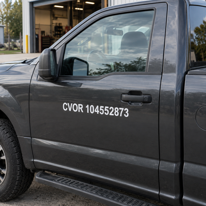 Solid white CVOR 104552873 vinyl lettering on the dark driver door of a plain work truck outside a workshop.