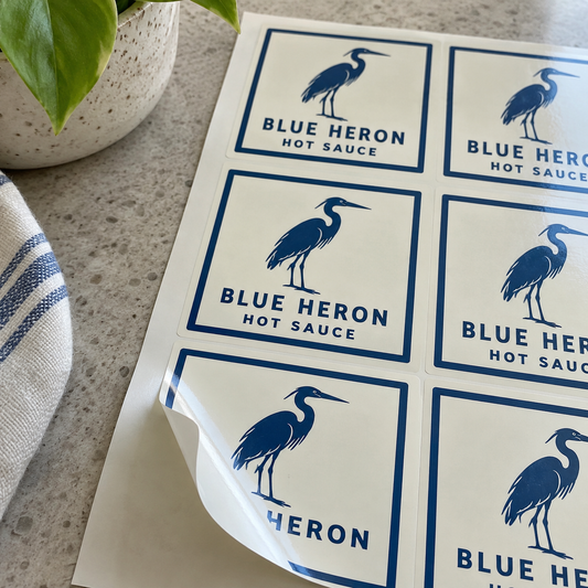 A sheet of Blue Heron Hot Sauce square labels with one corner lifted.