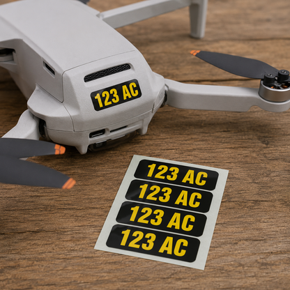Drone registration number decals in yellow on black vinyl reading 123 AC, one applied to a grey multirotor drone with a sheet of matching peel-and-stick decals on a wood surface
