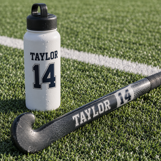 Taylor name and number 14 stickers on a field hockey stick and water bottle on turf.