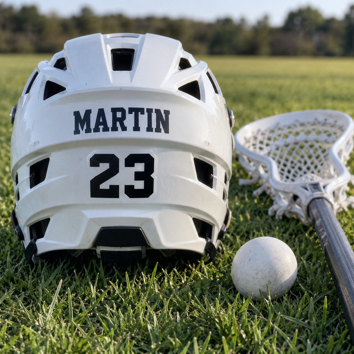 Martin name and number 23 stickers on a lacrosse helmet beside a stick and ball on grass.