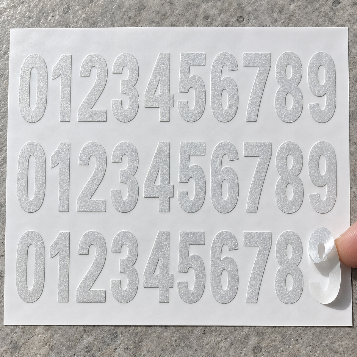 Three sets of reflective vinyl digits 0 to 9 on white backing paper, with one digit partially peeled by a fingertip.