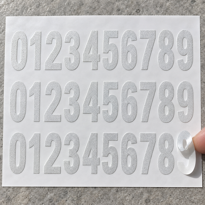 Three sets of reflective vinyl digits 0 to 9 on white backing paper, with one digit partially peeled by a fingertip.