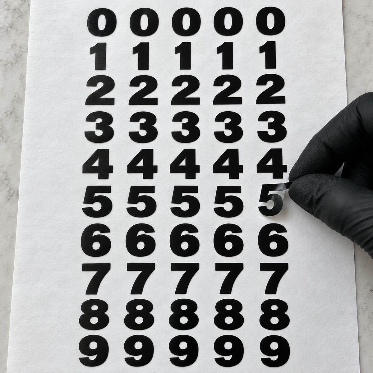 A black-gloved hand lifts one of five black vinyl 5s from white backing sheets arranged in ten rows of five identical digits, from 0 through 9.