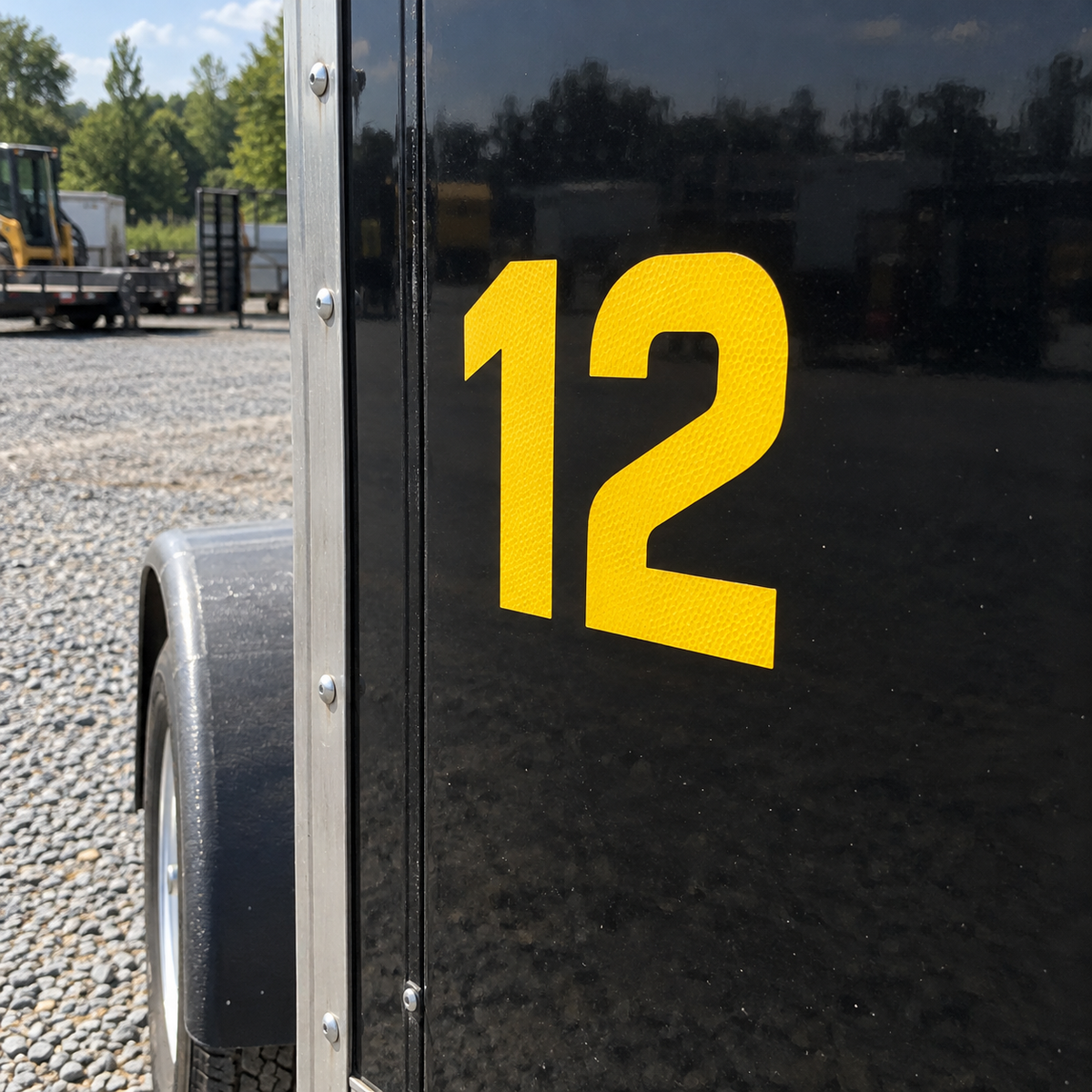 Yellow reflective vinyl number 12 on the side of a black enclosed cargo trailer in a gravel equipment yard.