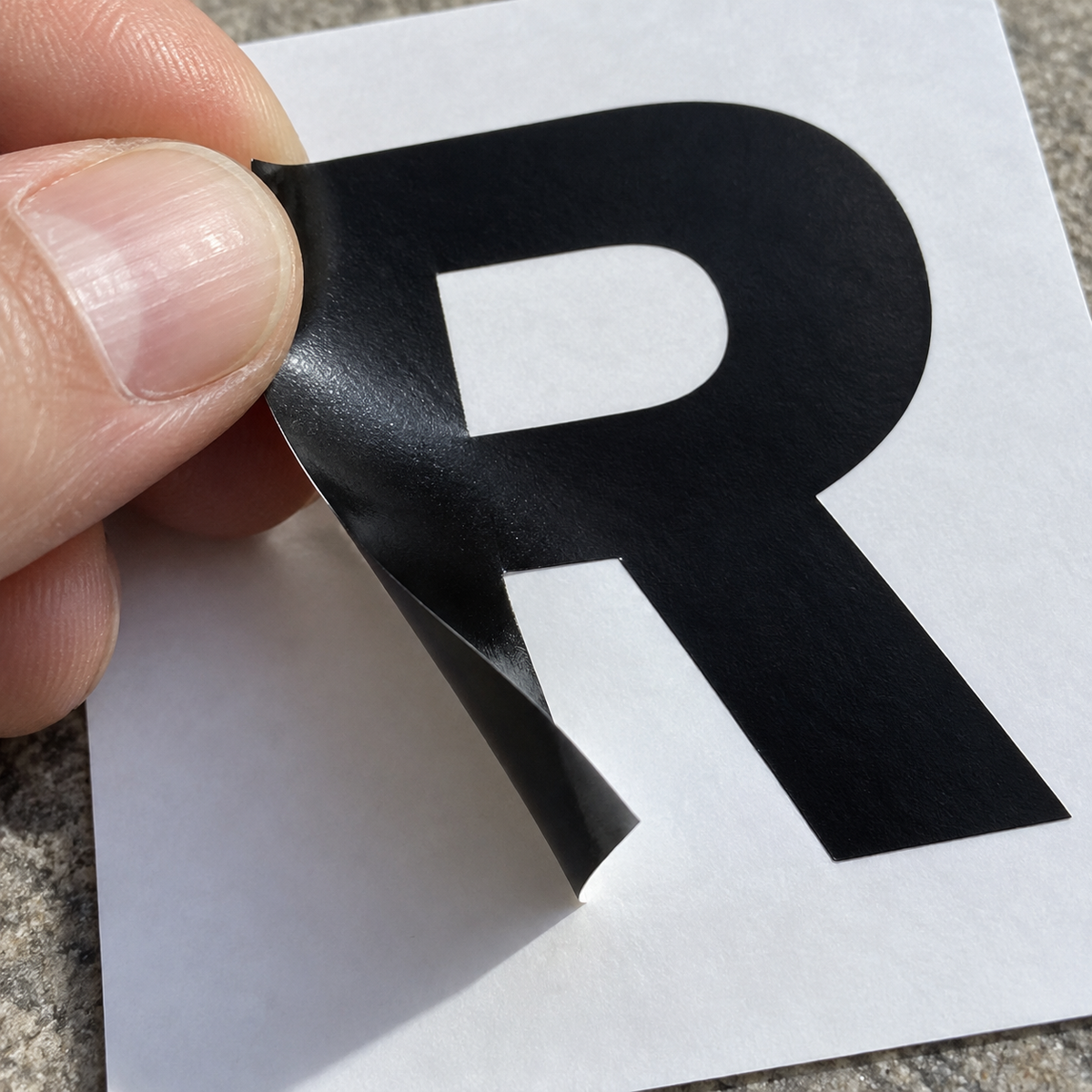 A hand peels a black vinyl capital R from its white backing sheet, showing crisp cut edges. - Vinyl Alphabet Letters — A-Z 0-9 Kit