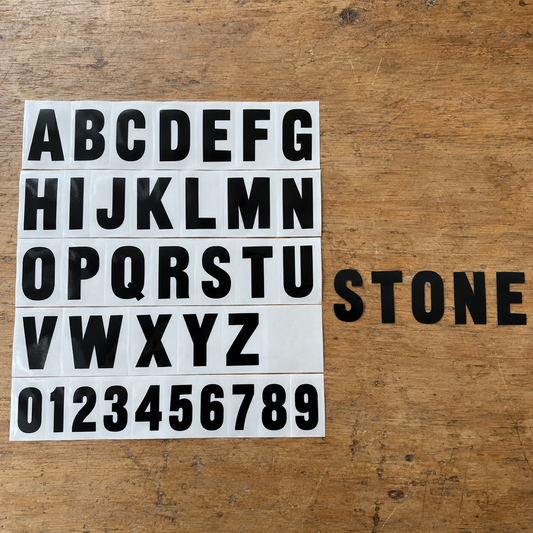 Black vinyl capital letters A to Z and numbers 0 to 9, each once on white backing sheets, beside five peeled letters spelling STONE on a wooden workbench. - Vinyl Alphabet Letters — A-Z 0-9 Kit