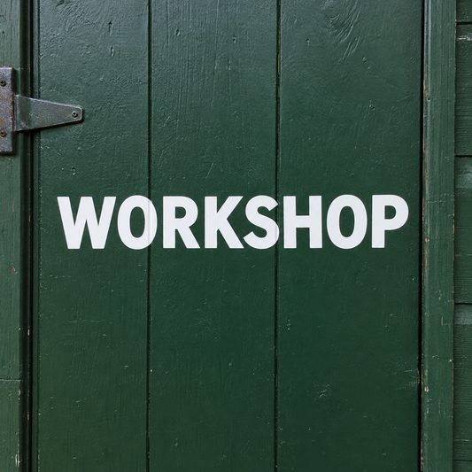 White vinyl capitals spelling WORKSHOP applied across a dark green painted shed door in daylight. - Vinyl Alphabet Letters — A-Z 0-9 Kit